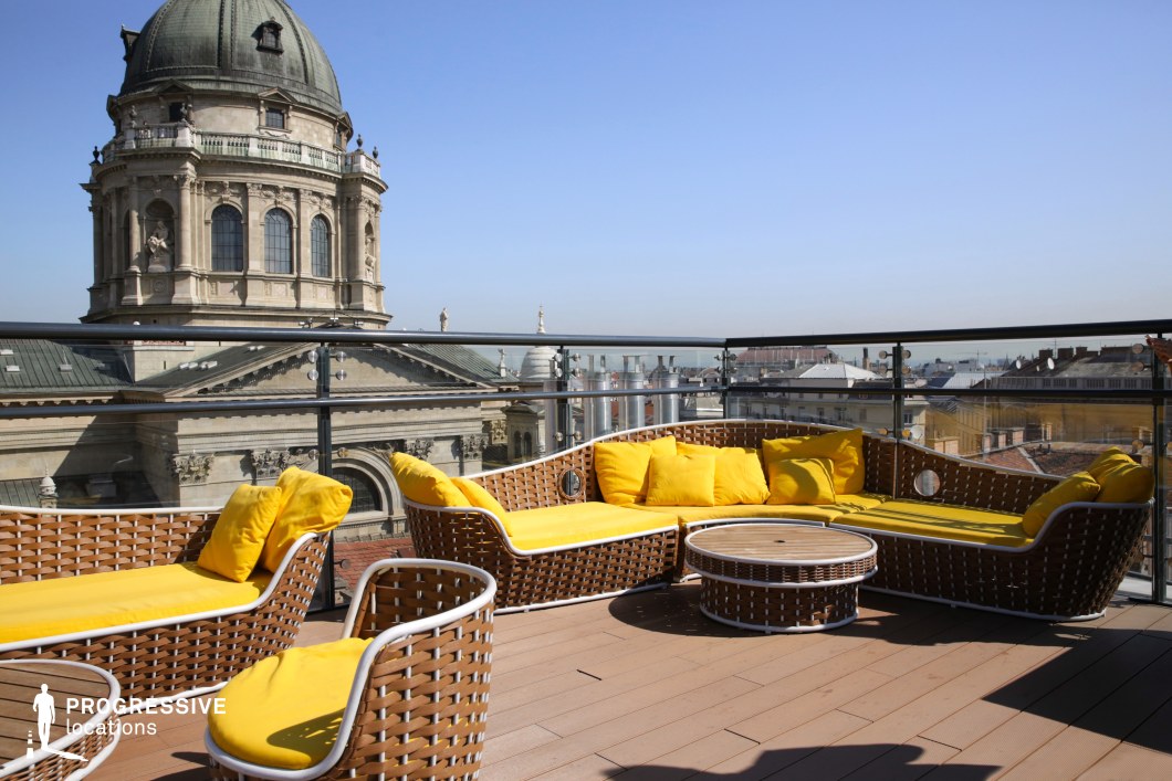 This stylish rooftop terrace features woven rattan furniture with bright yellow cushions, creating a lively contrast against the neutral-toned wooden decking. A sleek glass railing preserves the open, unobstructed view of the surrounding cityscape, dominated by an ornate domed historic building in the background. The combination of contemporary outdoor seating and classic architectural surroundings makes this an ideal filming location for high-end lifestyle scenes, social gatherings, or cinematic moments in romance and drama genres, emphasizing urban elegance and exclusivity.