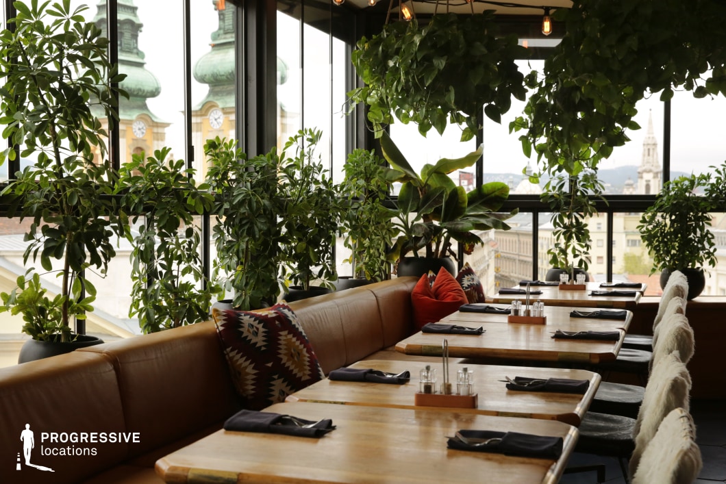 This rooftop restaurant blends natural elements with urban charm, featuring floor-to-ceiling windows that frame an impressive cityscape with historic architecture. The warm wooden tables, earthy leather seating, and plush cushions create a relaxed yet refined ambiance, while abundant greenery softens the modern interior. Suspended plants add depth and texture, enhancing the inviting atmosphere. The balance between rustic and contemporary aesthetics makes this an ideal filming location for romantic scenes, social gatherings, or lifestyle productions that emphasize intimate yet sophisticated settings.