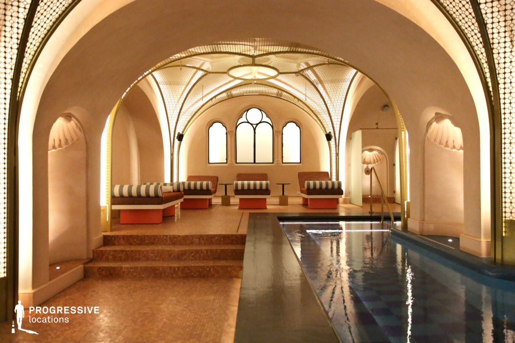 This luxurious spa blends historical architectural influences with contemporary elegance. The vaulted ceilings, adorned with intricate gold mesh detailing, create a grand yet intimate atmosphere. Soft, warm lighting accentuates the arched alcoves and shell-shaped wall niches, enhancing the space's serene ambiance. The relaxation area features plush, striped loungers set on elevated platforms, overlooking a sleek, reflective pool. With its balance of Moorish aesthetics and modern refinement, this location is ideal for high-end wellness retreats, cinematic settings, or editorial photo shoots emphasizing luxury and tranquility.