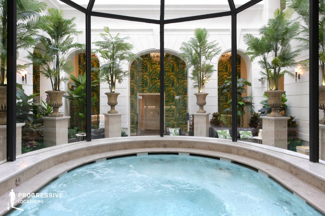 This elegant spa setting features a circular stone jacuzzi, framed by floor-to-ceiling glass windows that overlook a lush tropical courtyard. The space is designed with classical architectural elements, including arched white walls and stone pedestals supporting tall palm plants in ornate urns. The green and gold botanical wallpaper adds warmth and vibrancy, creating an immersive oasis-like atmosphere. Ideal for wellness retreats, luxury hospitality imagery, or high-end relaxation-themed productions, this space exudes tranquility and sophistication.