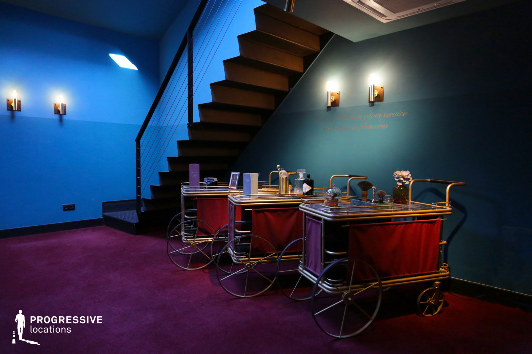 This striking interior blends deep blue and teal walls with a dramatic black staircase and plush red carpeting, creating a visually rich and atmospheric space. Gold-accented service carts with vintage-inspired designs add a touch of luxury, while soft, wall-mounted lighting enhances the moody ambiance. The interplay of contrasting colors and geometric elements makes this an ideal filming location for stylish noir, retro-futuristic narratives, or high-end dramas that require an intimate yet visually dynamic setting.