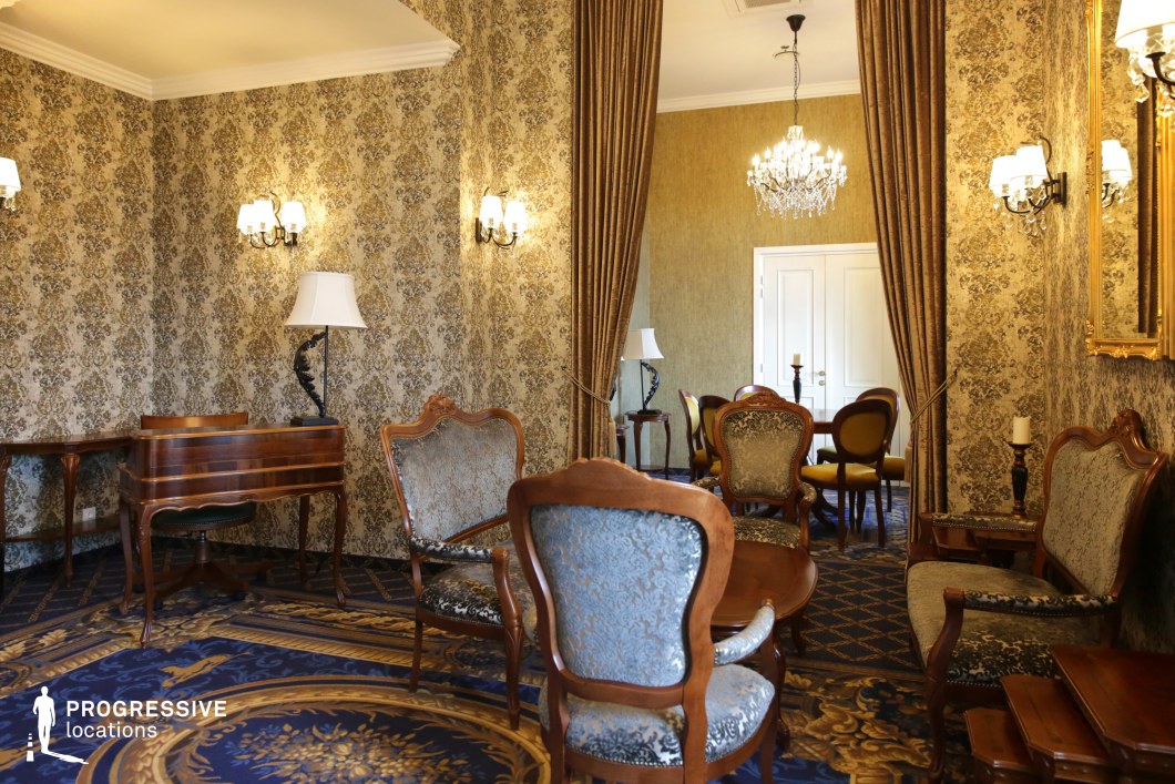 This refined vintage lounge features intricate wallpaper with a rich, classical pattern, complemented by deep blue and gold carpeting. The carved wooden furniture, upholstered in luxurious brocade, adds to the sophisticated ambiance, while soft lighting from wall sconces and chandeliers enhances the warm, intimate atmosphere. Heavy draped curtains separate the rooms, contributing to an air of exclusivity and privacy. This setting is ideal for period dramas, historical thrillers, or aristocratic intrigue, providing a visually detailed and atmospheric backdrop for refined conversations and high-society interactions.