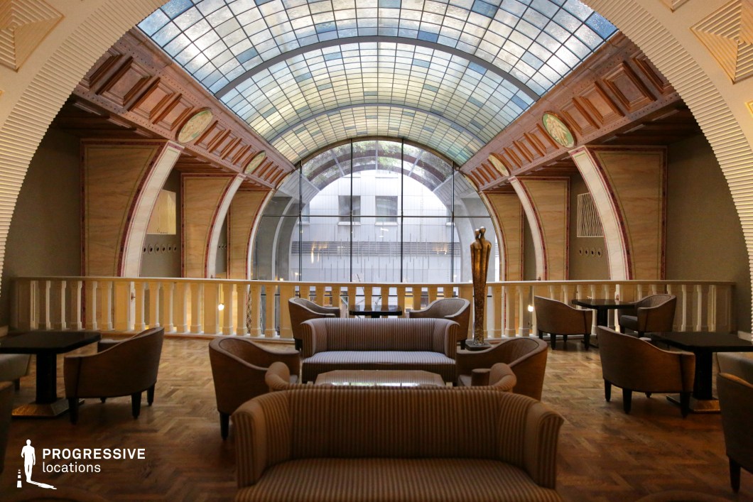 This refined lounge blends Art Deco and modernist elements, featuring a striking curved glass ceiling that allows soft, diffused light to filter into the space. The wooden paneling with intricate geometric detailing, along with warm-toned parquet flooring, creates an inviting yet sophisticated atmosphere. A minimalist bronze statue serves as a focal point, adding an artistic touch. The open balcony railing enhances the sense of spaciousness, while plush seating arrangements provide a luxurious setting. This location is ideal for stylish dramas, high-end espionage thrillers, or introspective character-driven films, offering a visually distinct and atmospheric backdrop.