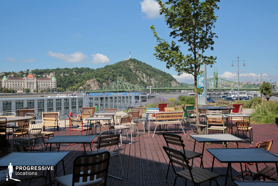 This open-air café terrace offers a stunning panoramic view of a historic European riverside, featuring a mix of wooden and metal-framed tables and chairs arranged informally on a dark wooden deck. The setting is complemented by lush greenery and a single tree, adding a natural touch to the relaxed urban atmosphere. In the background, an iconic green steel bridge spans the river, connecting the cityscape, while a grand classical-style building with distinctive towers enhances the architectural contrast. The hillside beyond, covered in dense foliage and crowned by a statue, adds depth to the composition, making this location ideal for capturing leisurely outdoor moments with a striking city-meets-nature backdrop.