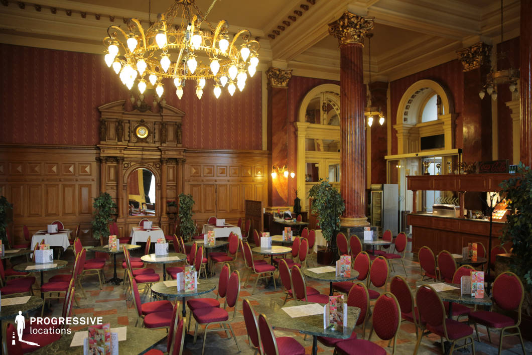 This opulent café exudes classic European elegance with its high ceilings, grand chandeliers, and richly decorated interiors. The space is defined by tall, marbled columns with gold-accented capitals, intricate wood paneling, and deep red wallpaper with traditional patterns, evoking a regal ambiance. The seating consists of round marble-topped tables surrounded by red upholstered chairs with gold trim, arranged in a spacious yet intimate layout. A central wooden clock and mirror add to the historic charm, while arched windows and doorways provide architectural depth. The polished stone floor reflects the warm lighting, enhancing the venue’s luxurious and timeless appeal, making it an ideal setting for refined social gatherings or period-style film scenes.