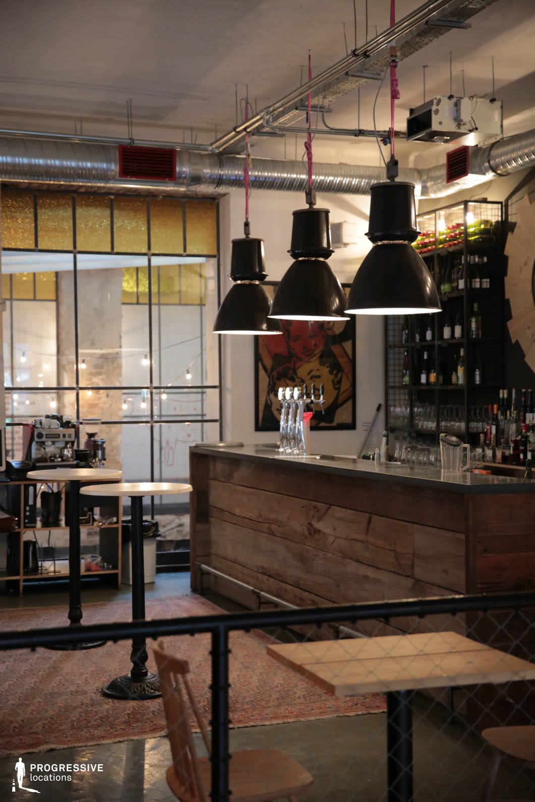 This contemporary bar combines industrial and rustic design elements, featuring a raw wooden bar counter with a sleek metal top, complemented by exposed ductwork and large black pendant lamps hanging from the ceiling. The open shelving behind the counter showcases an array of bottled drinks and glassware, enhancing the casual yet stylish atmosphere. A mix of high-top tables with minimalist wooden stools and black metal accents adds to the venue’s urban aesthetic. The background reveals large industrial windows with metal framing, allowing natural light to filter through, while vintage-style artwork and string lights contribute to the warm, eclectic ambiance. The combination of raw materials, muted tones, and soft lighting makes this space ideal for capturing scenes with an edgy, modern feel.