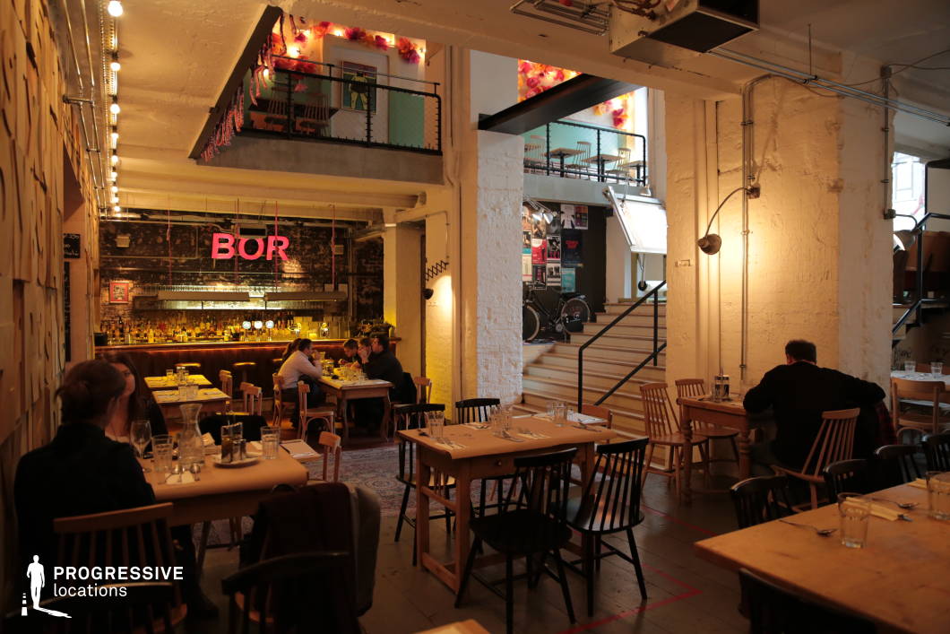 This stylish restaurant blends industrial and rustic design elements, creating a warm and inviting dining atmosphere. The space features exposed white brick walls, visible wiring, and raw metal accents, contrasted by wooden furniture and soft ambient lighting. A neon 