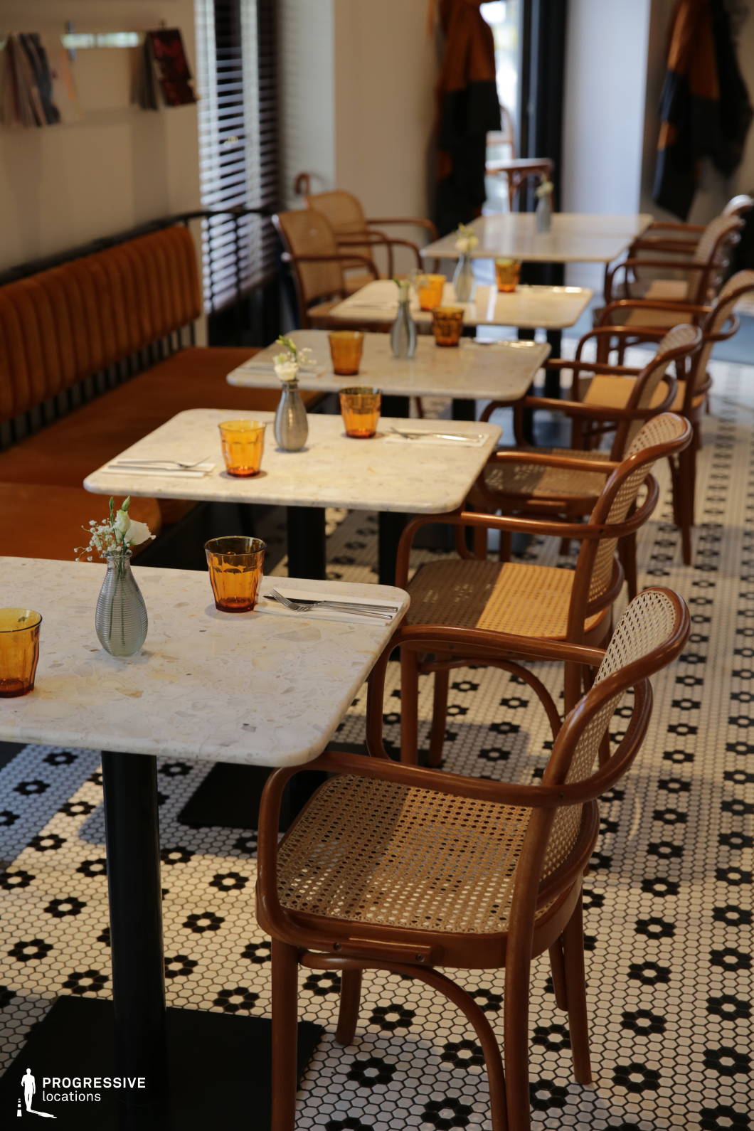 This stylish café blends classic and contemporary elements, featuring terrazzo marble tables, bentwood chairs with woven cane seats, and a vintage-inspired black-and-white hexagonal tile floor. The warm-toned leather banquette seating along the wall adds comfort, while minimalist décor, including small vases with fresh flowers and amber-colored glassware, enhances the inviting ambiance. Natural light filters through the space, highlighting the soft neutral palette and clean lines of the furniture. The refined yet casual setting, with neatly arranged place settings and thoughtfully curated design details, makes this an ideal backdrop for intimate conversations or relaxed dining in an effortlessly elegant atmosphere.