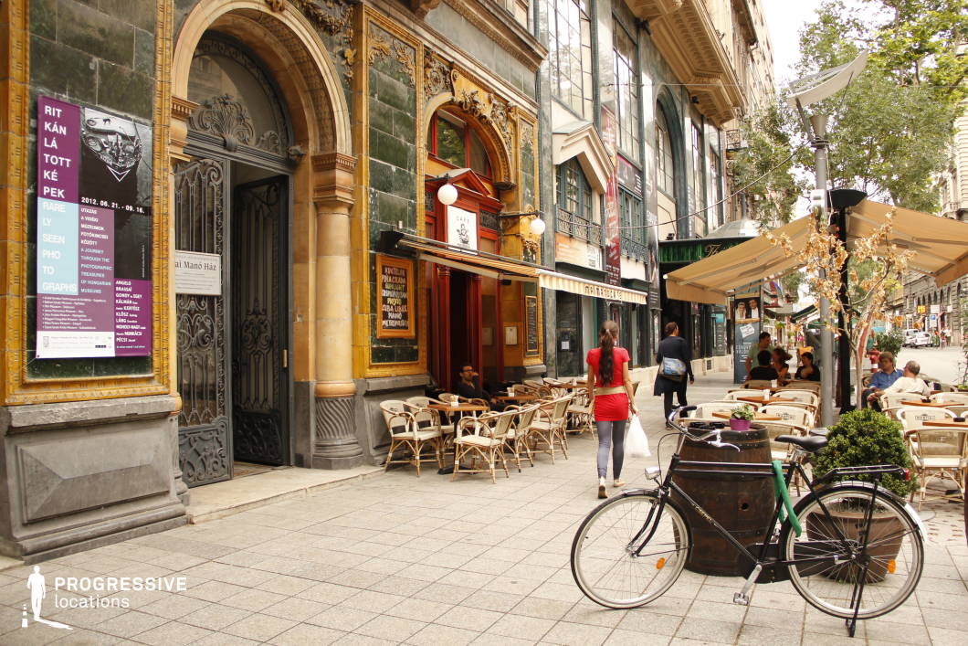 This Art Nouveau café, with its richly decorated façade, intricate ironwork, and ornate tilework, embodies early 20th-century European elegance, making it a visually compelling urban setting. The arched entrance and wooden accents enhance its historic appeal, while the outdoor terrace, furnished with rattan chairs and shaded by a retractable canopy, extends into a pedestrian-friendly street, creating a lively yet refined atmosphere. The cobblestone walkway and surrounding historic façades add depth and continuity to the streetscape, making this location ideal for sophisticated urban scenes, period pieces, or upscale dining visuals. The blend of decorative architectural elements with modern functionality provides a versatile backdrop for creative productions.