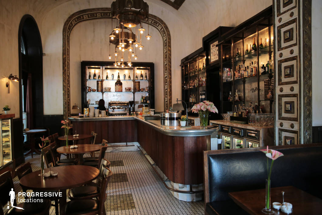 This Art Deco-style bar combines vintage elegance with modern functionality, featuring rich wooden paneling, intricate mosaic tile flooring, and a gracefully curved marble-topped bar as the focal point. The high ceiling, adorned with a statement chandelier of suspended glowing bulbs, enhances the grandeur, while geometric wall patterns and metallic accents reinforce the early 20th-century aesthetic. Illuminated glass shelves stocked with premium spirits create a sophisticated backdrop, complemented by round wooden tables and leather-upholstered seating that evoke a refined yet intimate atmosphere. The warm lighting and detailed ornamentation make this space ideal for upscale social scenes, period pieces, or atmospheric lounge settings.