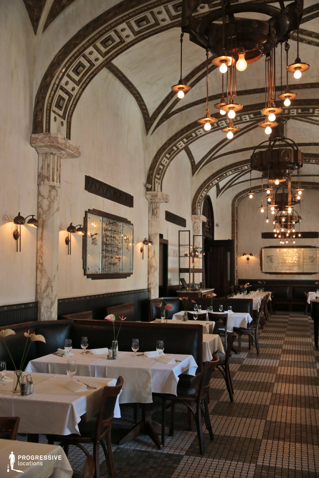 This elegantly designed Art Deco restaurant features grand vaulted ceilings adorned with intricate geometric patterns and marble-accented columns, creating a sense of historic refinement. The space is illuminated by suspended vintage chandeliers and warm wall sconces, enhancing the soft ambiance. The dark leather banquettes, neatly arranged tables draped in crisp white linens, and mosaic-tiled flooring contribute to an upscale yet intimate dining environment. The symmetrical layout and repeating arches establish a visually harmonious setting, making this location ideal for period settings, fine dining scenes, or stylish urban storytelling. The blend of luxurious materials and ambient lighting creates a timeless and cinematic atmosphere.