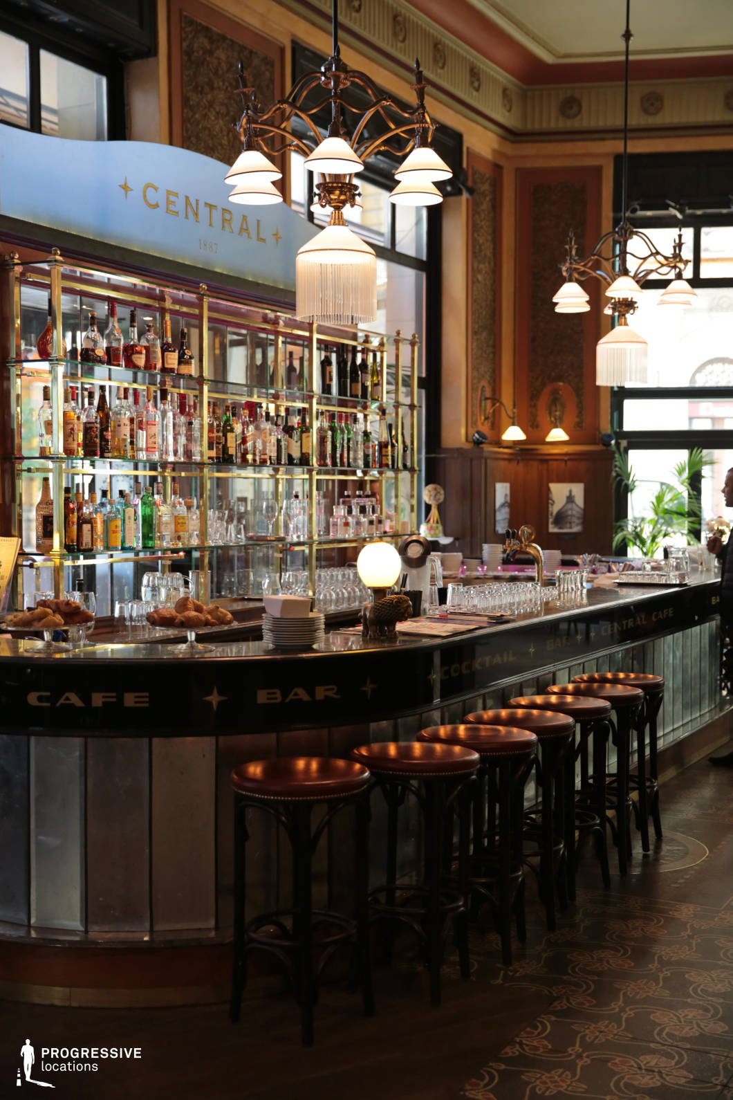 This sophisticated bar space exudes a vintage European charm, featuring an ornate dark counter with metallic accents and a row of classic leather-topped barstools that enhance its refined yet inviting ambiance. The mirrored back bar with brass shelving elegantly displays an extensive liquor selection, reflecting the warm lighting from the intricate chandeliers above. Rich wood paneling, decorative wall details, and large windows framed by dark trim contribute to the timeless elegance of the interior. The combination of polished materials, ambient lighting, and intricate Art Nouveau-inspired design elements creates an atmosphere that is both upscale and intimate, ideal for high-end social gatherings, period film settings, or refined cocktail experiences.