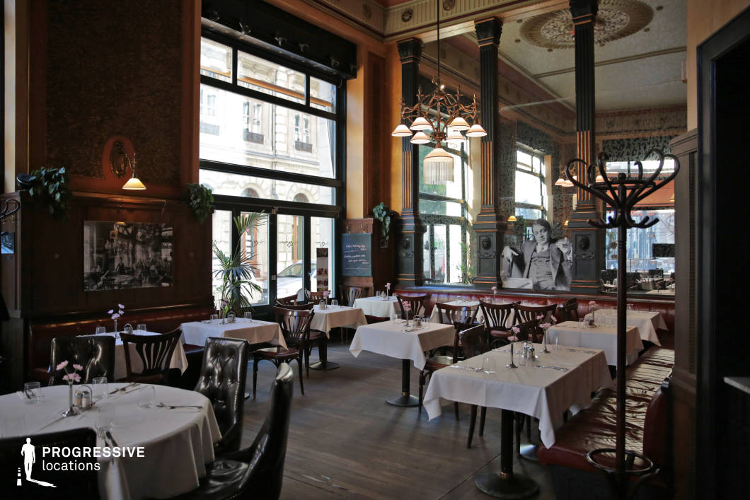 This classic café-restaurant exudes vintage charm with its high ceilings, intricate moldings, and large mirrors that enhance the sense of space and light. The dark wood paneling, ornate columns, and chandeliers contribute to an old-world European sophistication, while the floor-to-ceiling windows allow ample natural light to highlight the rich textures and details. White tablecloths, elegantly set tables, and a mix of tufted leather banquettes and wooden chairs create a refined yet welcoming dining atmosphere. The presence of historical photographs and decorative elements adds to the nostalgic ambiance, making this space ideal for period film settings, upscale dining experiences, or stylish social gatherings.