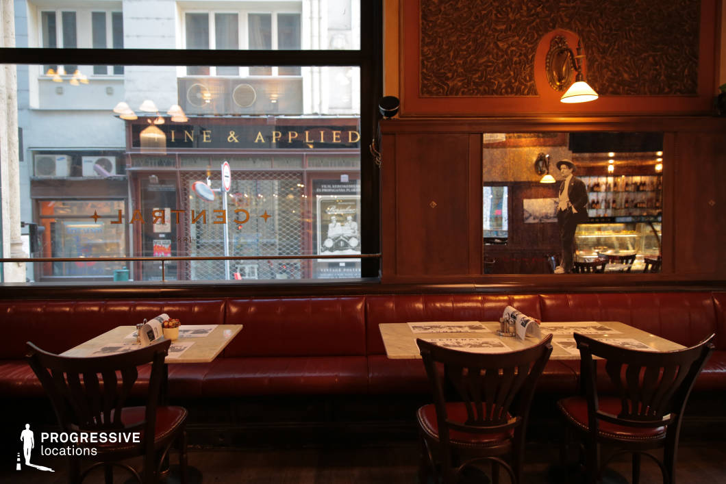 This intimate café space exudes a nostalgic, old-world atmosphere with its dark wood paneling, deep red leather banquettes, and vintage-inspired décor. The marble-topped tables and classic wooden chairs add to the refined yet casual setting, while the large window provides a glimpse of the street outside, subtly blending the café’s warm interior with the urban backdrop. A striking mirror installation reflects the space, enhancing its depth and ambiance, with artistic imagery adding a layer of character. The combination of soft wall lighting and ambient reflections creates a cozy, cinematic aesthetic, making it ideal for period settings, stylish social gatherings, or relaxed coffeehouse scenes.
