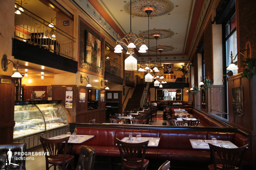 This opulent café-restaurant exudes classic European charm, featuring high ceilings adorned with intricate decorative moldings and grand chandeliers that cast a warm, inviting glow. The deep red leather banquettes and polished wooden furnishings create a rich, vintage aesthetic, further enhanced by framed historic photographs and ornamental wall sconces. Expansive windows flood the space with natural light, highlighting the spacious layout and mezzanine level, which adds depth and visual interest. A glass display case filled with pastries and desserts reinforces the café’s traditional elegance. The combination of period-style lighting, dark wood paneling, and refined décor makes this setting ideal for historical film productions, elegant dining experiences, or sophisticated social gatherings.