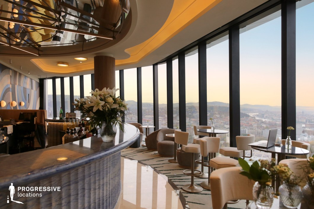 This luxurious sky lounge offers a breathtaking panoramic view of the city through its floor-to-ceiling windows, creating an open and airy ambiance. The interior design blends contemporary elegance with soft, organic elements, featuring curved furnishings, plush seating, and a statement bar with a sleek, marble-like finish. A mirrored ceiling with artistic lighting reflections adds depth and sophistication, complementing the curved architectural details. The neutral color palette, accented with warm lighting and subtle decorative touches like floral arrangements, enhances the refined atmosphere. Ideal for upscale social events, high-end dining, or cinematic urban settings, this space seamlessly combines modern luxury with an unparalleled cityscape backdrop.