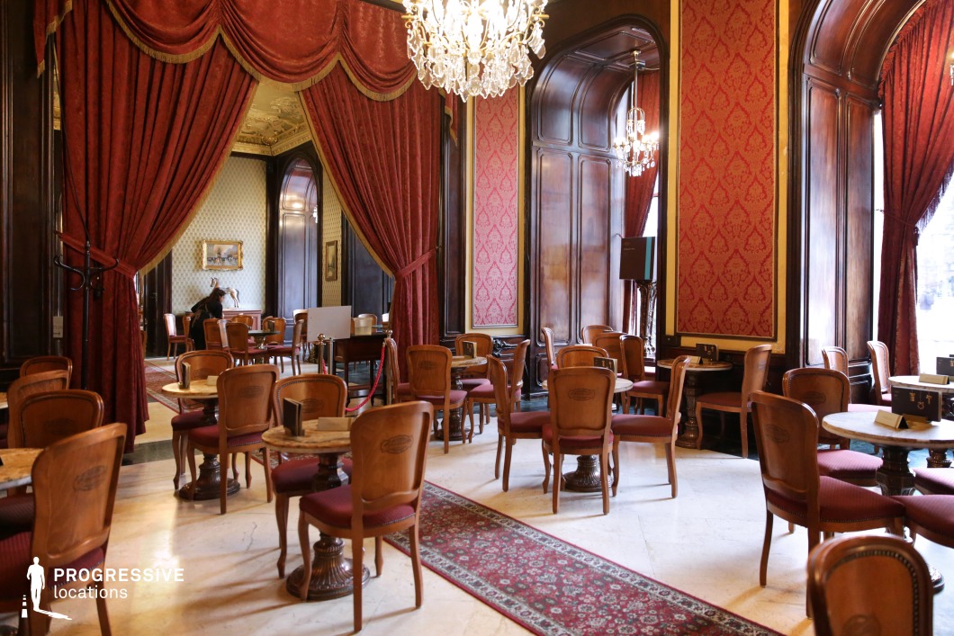 This opulent café interior embodies the grandeur of a bygone era, featuring high ceilings, crystal chandeliers, and rich wooden paneling that exude refined sophistication. The deep red damask-patterned walls and heavy draped curtains add warmth and a regal ambiance, complementing the plush upholstered chairs and intricately carved wooden tables. The marble flooring and elegant Persian-style rugs enhance the sense of luxury, while the arched windows allow natural light to illuminate the richly decorated space. The café’s timeless aesthetic makes it an ideal setting for period film productions, high-end social gatherings, or elegant dining experiences steeped in historical charm.