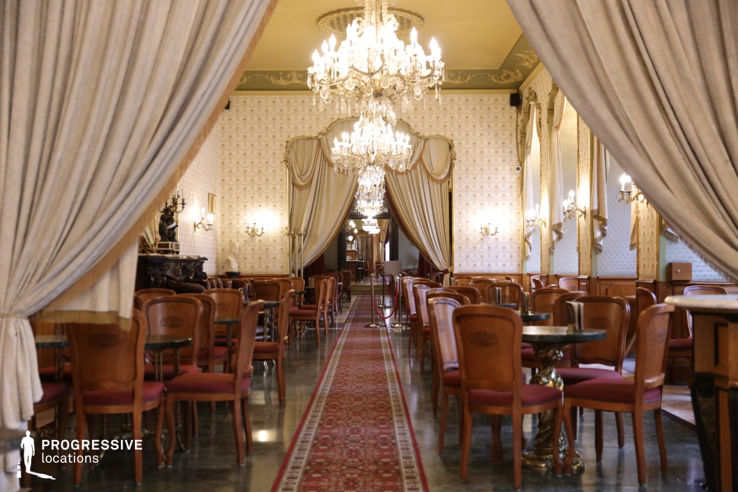 This grand café hall exudes opulence with its high ceilings, elaborate chandeliers, and luxurious drapery that frame the space in a regal ambiance. The polished marble flooring and intricate Persian-style carpet running down the central aisle add a refined touch, while the soft golden wallpaper and warm wooden paneling enhance the classic European aesthetic. The carefully arranged wooden chairs and round tables with sculpted legs create an inviting yet formal atmosphere. Ornate sconces and decorative elements, including statues and carved furnishings, contribute to the historical charm. This setting is ideal for period film productions, high-end social events, or an immersive fine dining experience in a timeless, aristocratic atmosphere.