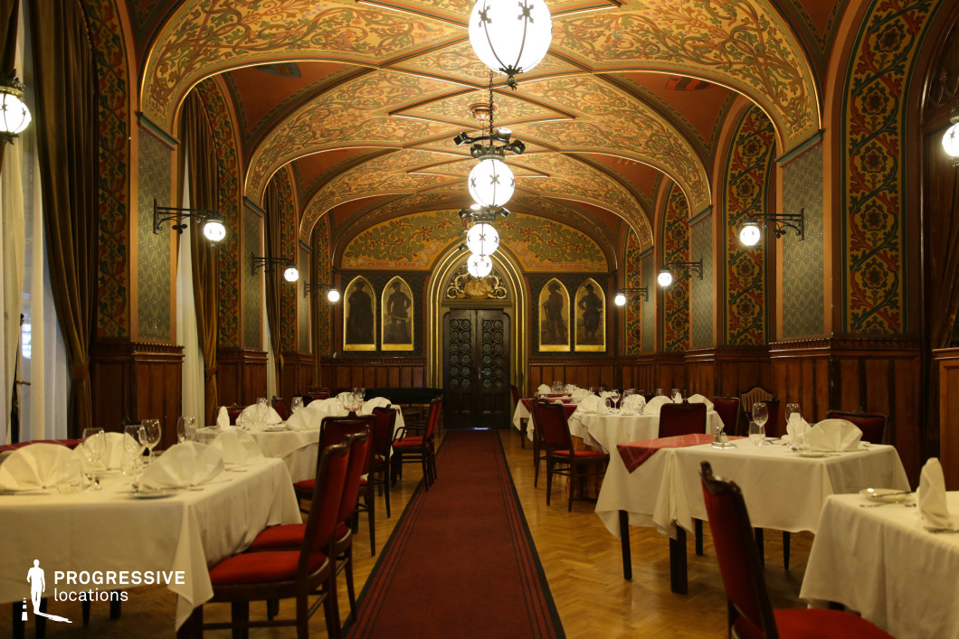 This breathtaking dining hall showcases a rich Gothic-inspired design, featuring an elaborately painted vaulted ceiling adorned with intricate floral and geometric patterns. The arched structure is emphasized by warm wooden paneling and deep red accents, creating a regal and intimate atmosphere. Rows of elegantly set tables with crisp white linens contrast with the dark wooden furnishings, while ambient globe lighting enhances the historic charm. The end of the hall is framed by a grand double-door entrance, flanked by medieval-style frescoes, adding to the grandeur of the space. Ideal for period film settings, fine dining experiences, or high-end historical-themed events, this setting exudes an air of timeless elegance and aristocratic refinement.