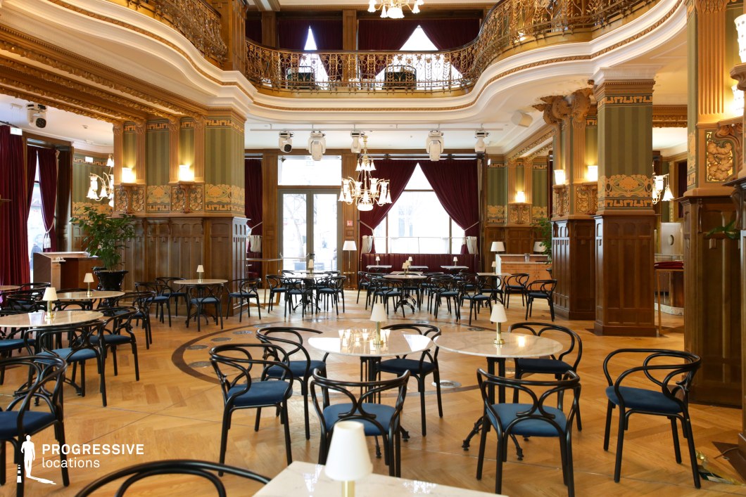 This spacious café showcases exquisite Art Nouveau architecture, featuring intricate gold-accented wood paneling, sculpted columns, and an elegant mezzanine with ornate wrought-iron railings. The high ceilings, adorned with decorative moldings and chandeliers, enhance the grandeur of the space, while large windows framed by rich burgundy drapes allow natural light to highlight the warm parquet flooring. The interior is furnished with round marble-topped tables and classic black bentwood chairs, creating a refined yet inviting atmosphere. Ideal for high-end dining, period film settings, or sophisticated social gatherings, this venue seamlessly blends historic charm with timeless elegance.