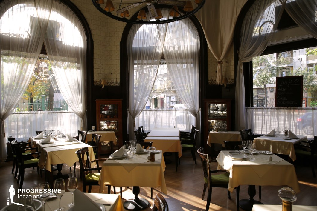 This refined café exudes a timeless European elegance, featuring tall arched windows draped with sheer white curtains that softly filter natural light, creating a warm and inviting ambiance. The interior blends vintage and sophisticated elements, with dark wooden furnishings, polished floors, and neatly arranged tables adorned with crisp yellow tablecloths and fine dining settings. Antique display cabinets and subtle decorative details add character, while the large windows connect the cozy atmosphere with the surrounding cityscape. Ideal for intimate dining, elegant gatherings, or period film settings, this café offers a perfect balance of classic charm and airy sophistication.
