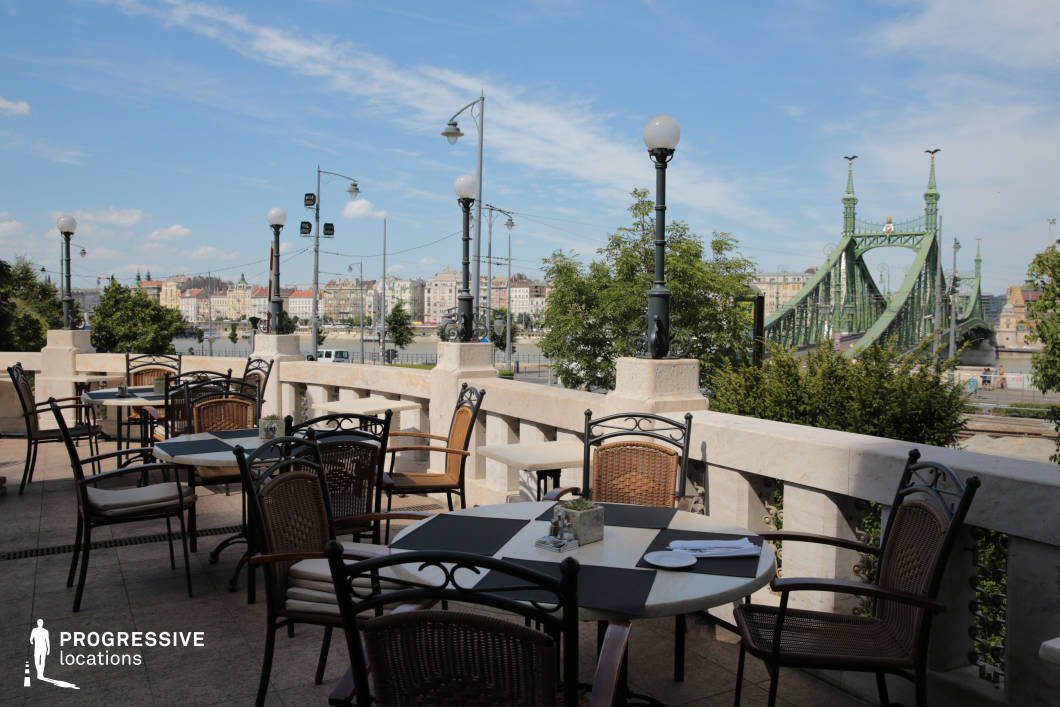 This elegant outdoor dining terrace offers a picturesque setting with a sweeping view of the river, historic architecture, and a striking green bridge in the background. The classic stone balustrade, adorned with vintage-style lampposts, enhances the refined ambiance, while the mix of wrought iron and wicker chairs provides a balance of comfort and sophistication. The neatly arranged tables with subtle décor create an inviting yet upscale dining experience. Surrounded by lush greenery and open skies, this terrace is an ideal setting for relaxed yet stylish gatherings, high-end dining, or cinematic scenes capturing the charm of a European riverside café.