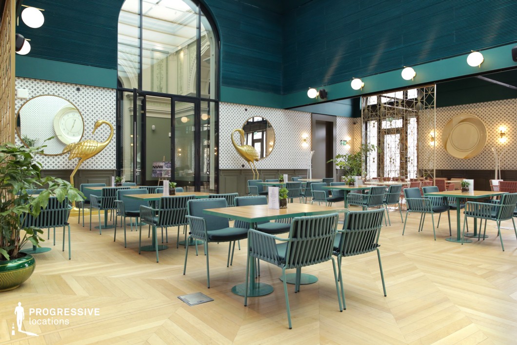 This stylish café blends modern elegance with Art Deco-inspired elements, featuring a striking color palette of deep teal, gold accents, and warm wood tones. The open, airy space is framed by high ceilings and large arched windows, allowing natural light to enhance the geometric patterns and reflective surfaces. Unique decorative elements, such as gold flamingo sculptures and circular wall mirrors, add a playful yet sophisticated touch. The seating consists of sleek, woven-back chairs and minimalist wooden tables, maintaining a balance of comfort and refinement. With its fusion of classic and contemporary design, this venue is ideal for upscale dining, stylish social gatherings, or visually striking film settings.