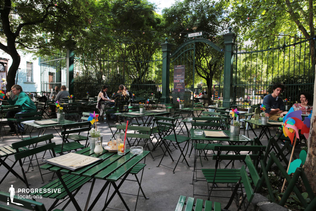 This inviting outdoor café is nestled within a lush green park, offering a tranquil retreat surrounded by towering trees and a decorative wrought-iron fence. The setting features classic green metal bistro tables and foldable chairs, creating a casual and communal atmosphere. Cheerful pinwheels and small potted plants add a playful, whimsical touch, enhancing the café’s vibrant and welcoming appeal. String lights draped across the space contribute to a cozy, intimate ambiance, making it an ideal location for relaxed social gatherings, leisurely afternoons, or film scenes that capture the charm of a laid-back European garden café.