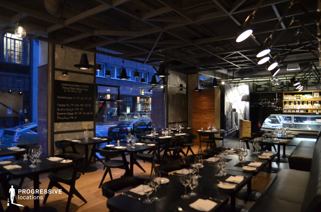 This sophisticated steakhouse exudes a modern industrial aesthetic, featuring dark minimalist furnishings, exposed ceiling beams, and raw concrete walls that create a bold yet refined atmosphere. Black tables and stools contrast with the warm wood flooring, while strategically placed lighting fixtures cast a moody ambiance. Floor-to-ceiling windows connect the space to the urban streetscape, adding an open and dynamic element. A chalkboard menu and delicatessen counter further enhance the upscale yet approachable dining experience. Ideal for high-end culinary experiences, stylish nightlife settings, or cinematic urban scenes, this venue blends industrial rawness with contemporary sophistication.