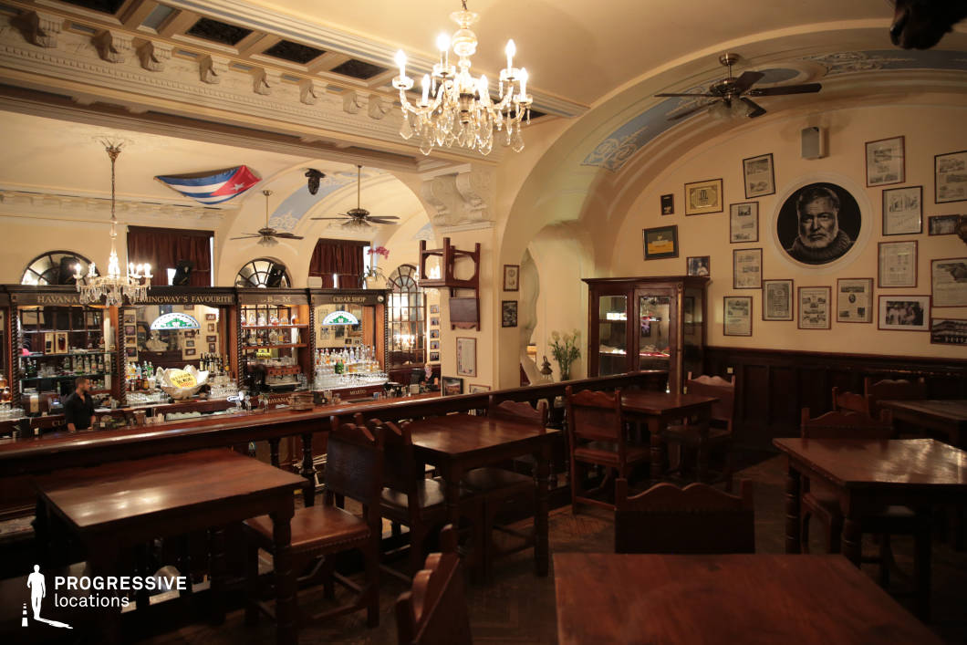 This historic Cuban bar exudes a warm, nostalgic ambiance with its dark wooden furnishings, ornate ceiling details, and vintage chandeliers casting a soft glow. The architectural elements, including elegant arches, decorative moldings, and stained-glass accents, reflect a colonial-era aesthetic, while the walls, adorned with framed memorabilia and a large portrait, add character and authenticity. The central wooden bar, with its mirrored back and extensive liquor display, serves as the focal point, complemented by an elevated seating area that introduces depth to the space. Ceiling fans and rich textures enhance the inviting atmosphere, making this location ideal for scenes requiring a timeless, old-world charm.