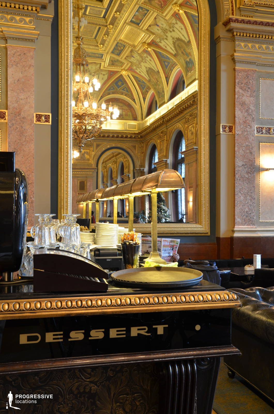 This luxurious café interior exudes historic grandeur with its gilded ceiling, intricate frescoes, and elaborate moldings that create a rich classical ambiance. The large arched windows framed by gold-trimmed columns introduce natural light, enhancing the polished marble and wood-paneled walls. A massive mirror amplifies the space, reflecting the ornate chandeliers and reinforcing the venue’s opulence. The foreground features a finely detailed counter marked 
