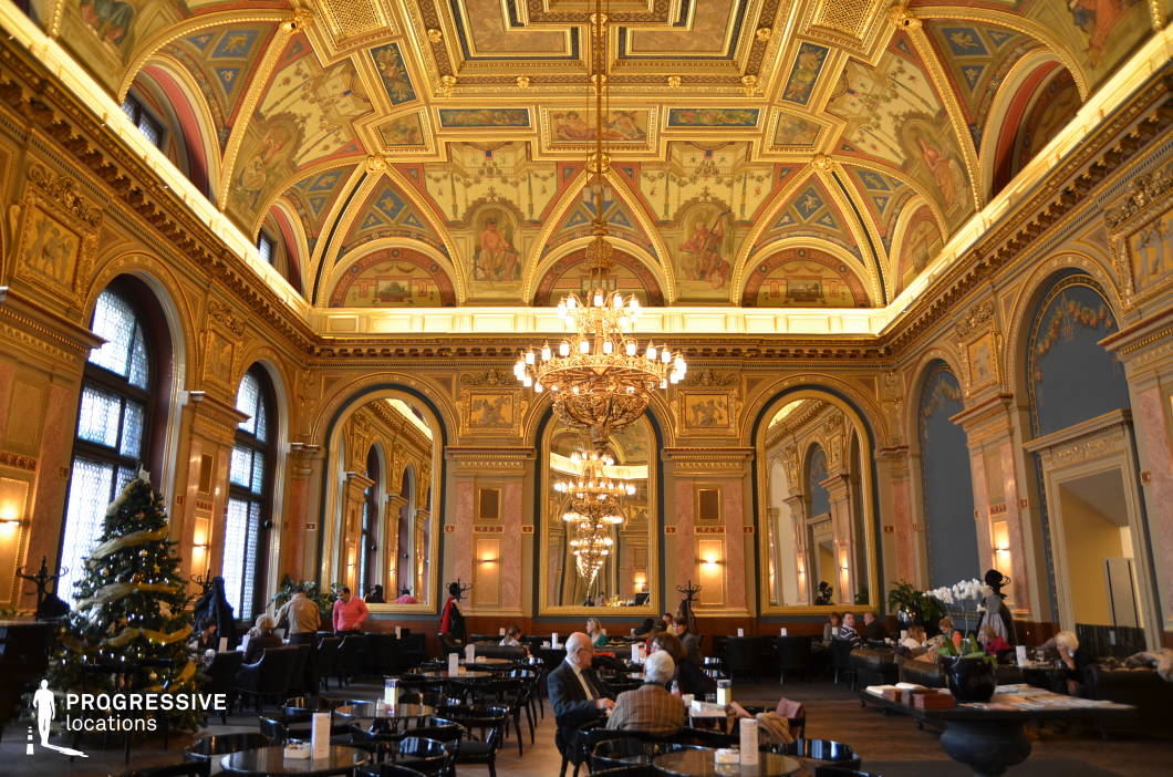 This opulent café hall embodies Renaissance elegance with its intricately decorated ceiling featuring frescoes, gold moldings, and elaborate patterns. Large arched windows with textured glass allow natural light to illuminate the pastel-toned walls adorned with gilded reliefs and classical motifs. Massive gold-framed mirrors enhance the sense of depth and symmetry, reflecting the warm glow of ornate chandeliers that add to the luxurious ambiance. The polished marble, wood paneling, and deep blue wall accents create a refined yet inviting atmosphere, while the sleek black furniture and plush seating provide a sophisticated contrast, making this an ideal setting for period-themed visuals or high-end dining experiences.