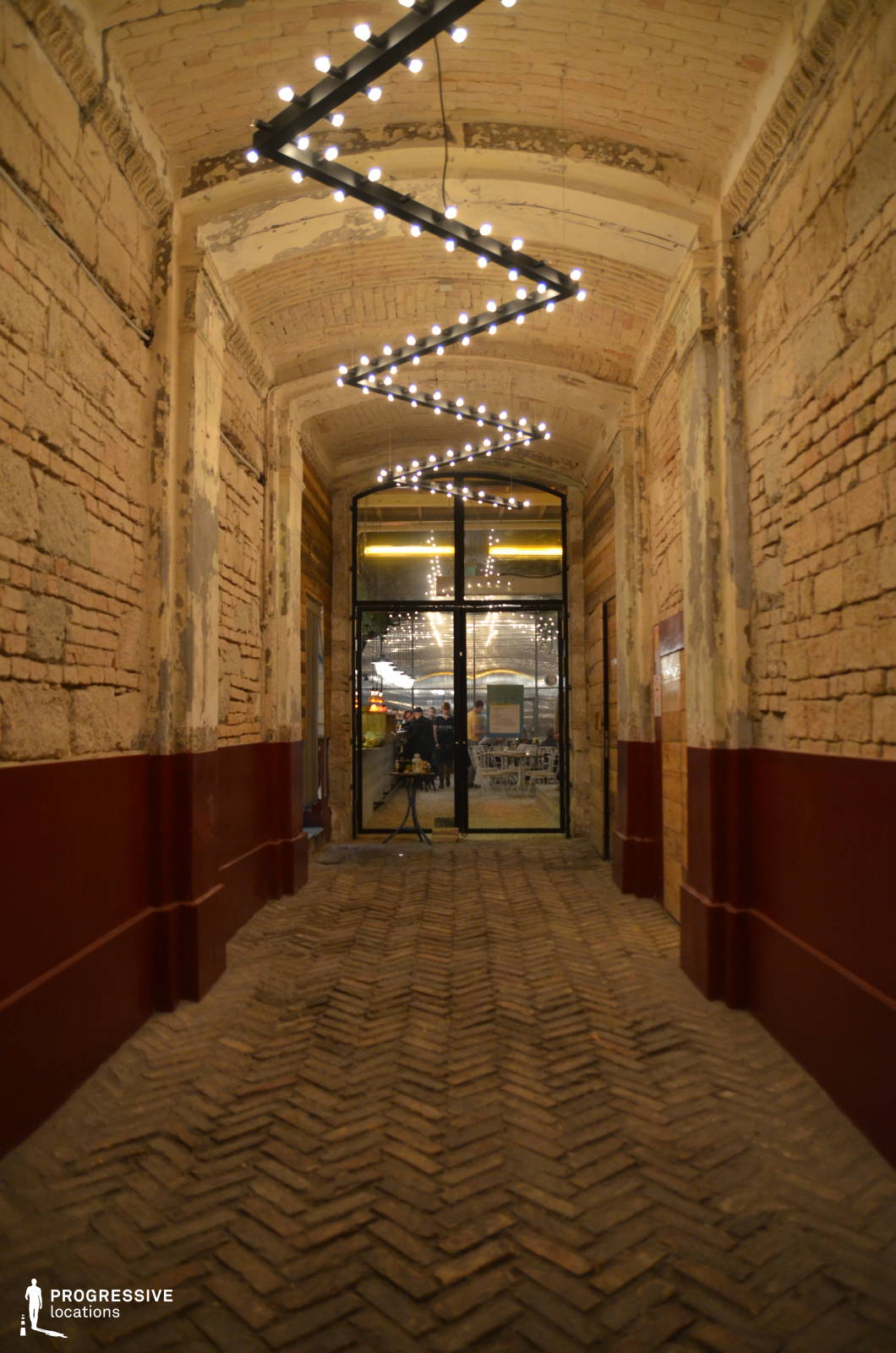 This rustic brick archway entrance blends historic charm with modern industrial design, featuring exposed aged brick walls, a vaulted ceiling, and a herringbone-patterned brick floor. The raw, unfinished textures of the walls contrast with the sleek black-framed glass door at the end of the corridor, which offers a glimpse into a warmly lit interior space. A striking geometric light installation zigzags along the ceiling, casting a contemporary glow that enhances the tunnel-like perspective. The combination of weathered materials and modern lighting elements creates a visually dynamic setting, ideal for urban nightlife, industrial-chic aesthetics, or moody, cinematic compositions.