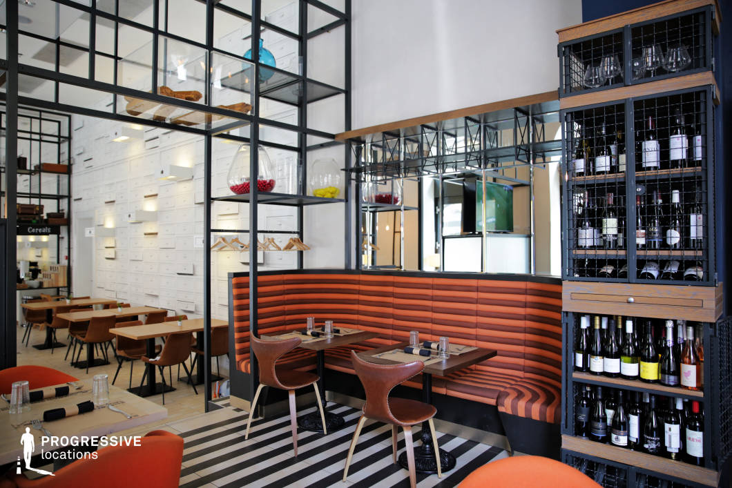 This stylish bistro combines industrial-chic elements with warm, modern accents, featuring an open layout with metal-framed shelving, geometric mirrors, and a bold black-and-white striped floor. The seating includes a mix of plush orange banquettes and sleek leather chairs, adding warmth to the structured aesthetic. A feature wall with a grid of white drawers creates a striking visual texture, while minimalist pendant lighting enhances the clean, modern ambiance. The wine display, encased in metal and wood, adds a refined touch, complementing the restaurant's contemporary yet inviting atmosphere, making it an ideal setting for casual fine dining or design-forward hospitality concepts.