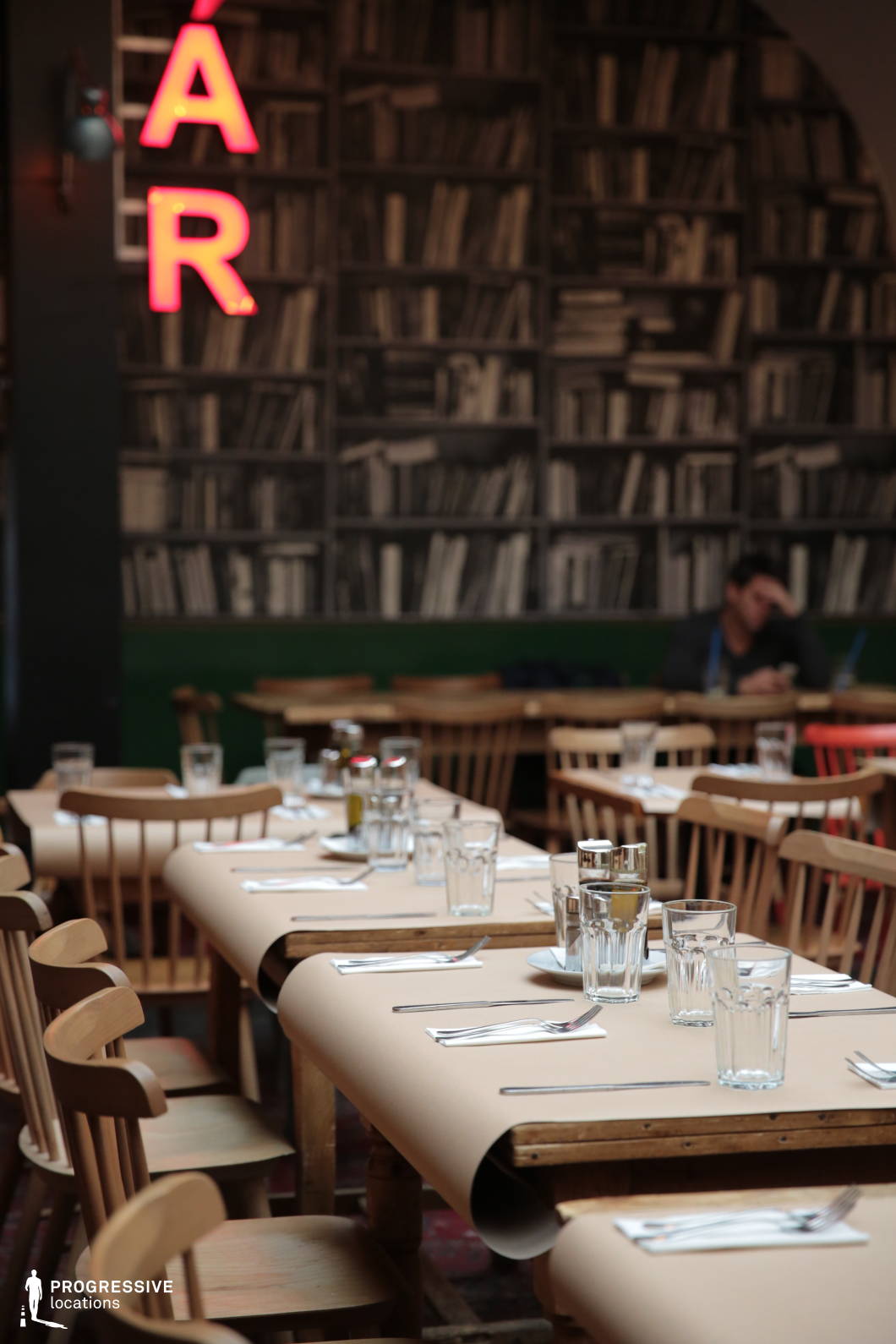 This intimate bistro combines a warm, rustic ambiance with a unique literary theme, featuring wooden communal tables covered in parchment-style paper, simple wooden chairs, and a striking black-and-white bookshelf mural as a backdrop. A red neon 