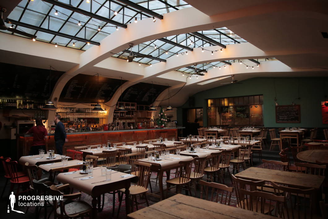 This spacious bistro blends rustic charm with industrial elements, featuring a striking glass ceiling supported by curved white beams, allowing natural light to illuminate the open dining area. The long wooden bar, with its distressed finish and overhead chalkboard menus, adds a vintage touch, while warm string lights create a cozy ambiance. The mix of wooden tables, mismatched chairs, and parchment-covered surfaces enhances the relaxed, communal atmosphere, complemented by Persian rugs and soft pendant lighting. The combination of raw materials, soft lighting, and an open-concept layout makes this an ideal setting for casual dining, social gatherings, or creative urban spaces.