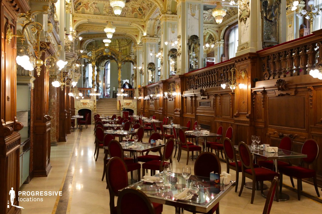 This opulent café hall showcases an exquisite Baroque-style interior with soaring gilded ceilings, intricate frescoes, and lavish chandeliers that cast a warm, golden glow. Rich wooden paneling with elaborate carvings lines the walls, complemented by ornate brass sconces and sculptural details that enhance the grandeur of the space. The seating arrangement features plush red velvet chairs paired with sleek glass tables, creating an elegant contrast between classic and modern elements. A dramatic staircase in the background leads to an upper level, framed by arched mirrors and gilded balustrades, further emphasizing the café’s palatial ambiance. This luxurious setting is ideal for high-end dining, period-piece visuals, or cinematic scenes requiring a majestic, old-world atmosphere.
