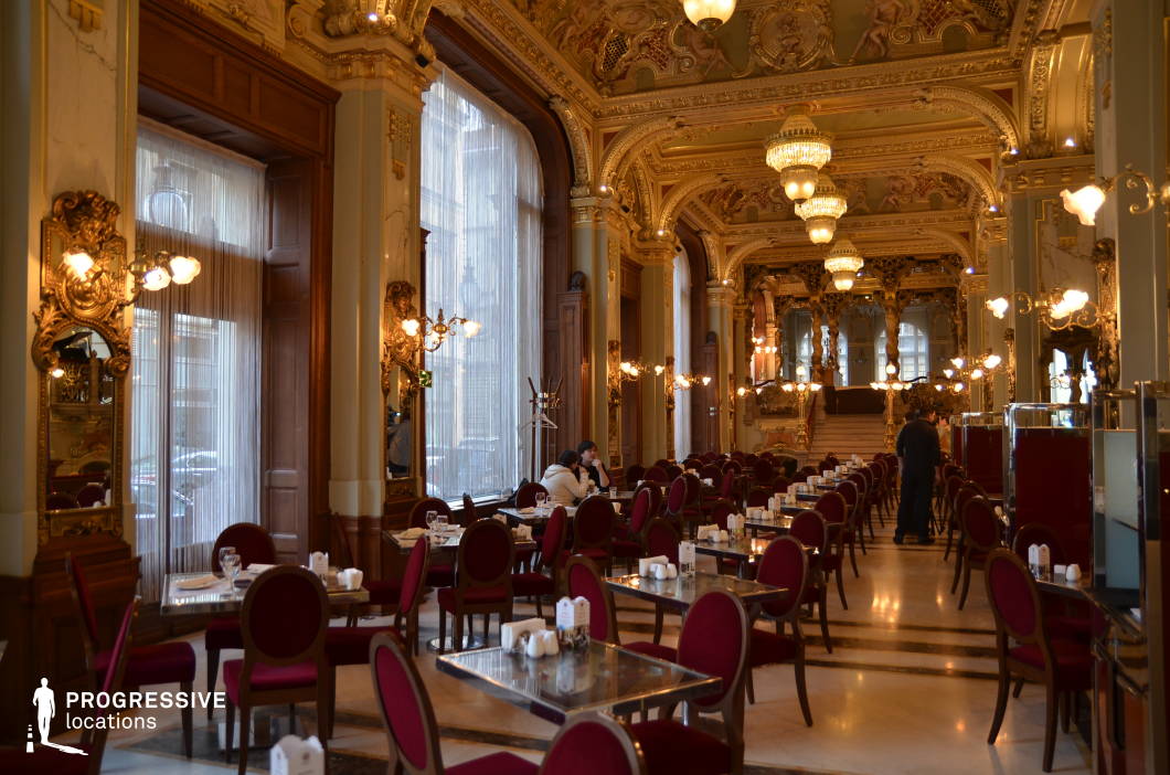 This grand café interior showcases an opulent Baroque-style design, featuring gilded ceilings adorned with intricate frescoes, massive chandeliers, and sculptural gold embellishments. Tall arched windows, framed by elegant drapery, allow natural light to enhance the richness of the warm wooden paneling and ornate wall mirrors. The seating arrangement includes plush red velvet chairs paired with reflective glass tables, creating a luxurious yet refined atmosphere. Elaborate gold-accented balustrades and a sweeping staircase in the background add a sense of depth and grandeur, making this space ideal for capturing a majestic, historic setting perfect for high-end dining or period-themed productions.