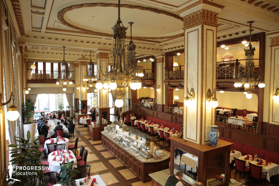 This grand Art Nouveau-style restaurant features an open, multi-level design with ornate architectural details, including high ceilings adorned with intricate moldings and elegant chandeliers suspended from decorative beams. The interior is defined by rich wooden paneling, marble-clad columns, and gilded accents, creating a refined and sophisticated atmosphere. A central buffet counter with a polished stone top serves as a focal point, surrounded by red-upholstered banquettes and neatly arranged white-tableclothed dining tables. Large windows framed by luxurious drapery allow natural light to enhance the space, while upper-level balconies with decorative railings add depth and grandeur. This stylish setting is ideal for upscale dining, formal gatherings, or period-inspired cinematic visuals.