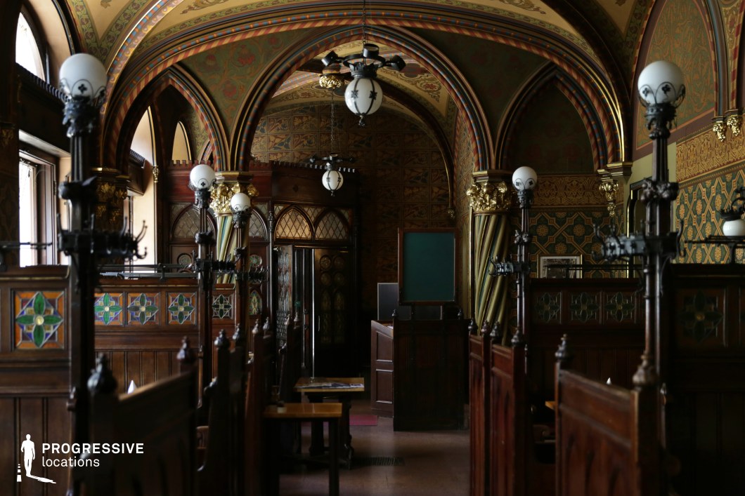 This grand historic dining hall features a richly decorated Gothic-inspired interior, characterized by pointed archways, dark wooden paneling, and intricate stained-glass inlays. The dimly lit space is lined with private wooden booths adorned with cast-iron light fixtures, creating an intimate and atmospheric setting. The vaulted ceiling and walls are embellished with elaborate patterns, gold detailing, and deep jewel-toned colors, adding to the venue’s old-world charm. Decorative doors with leaded glass panels and elegant drapery frame the background, enhancing the sense of grandeur and heritage. This striking location is ideal for period-themed visuals, classic European settings, or cinematic productions requiring a refined yet dramatic ambiance.