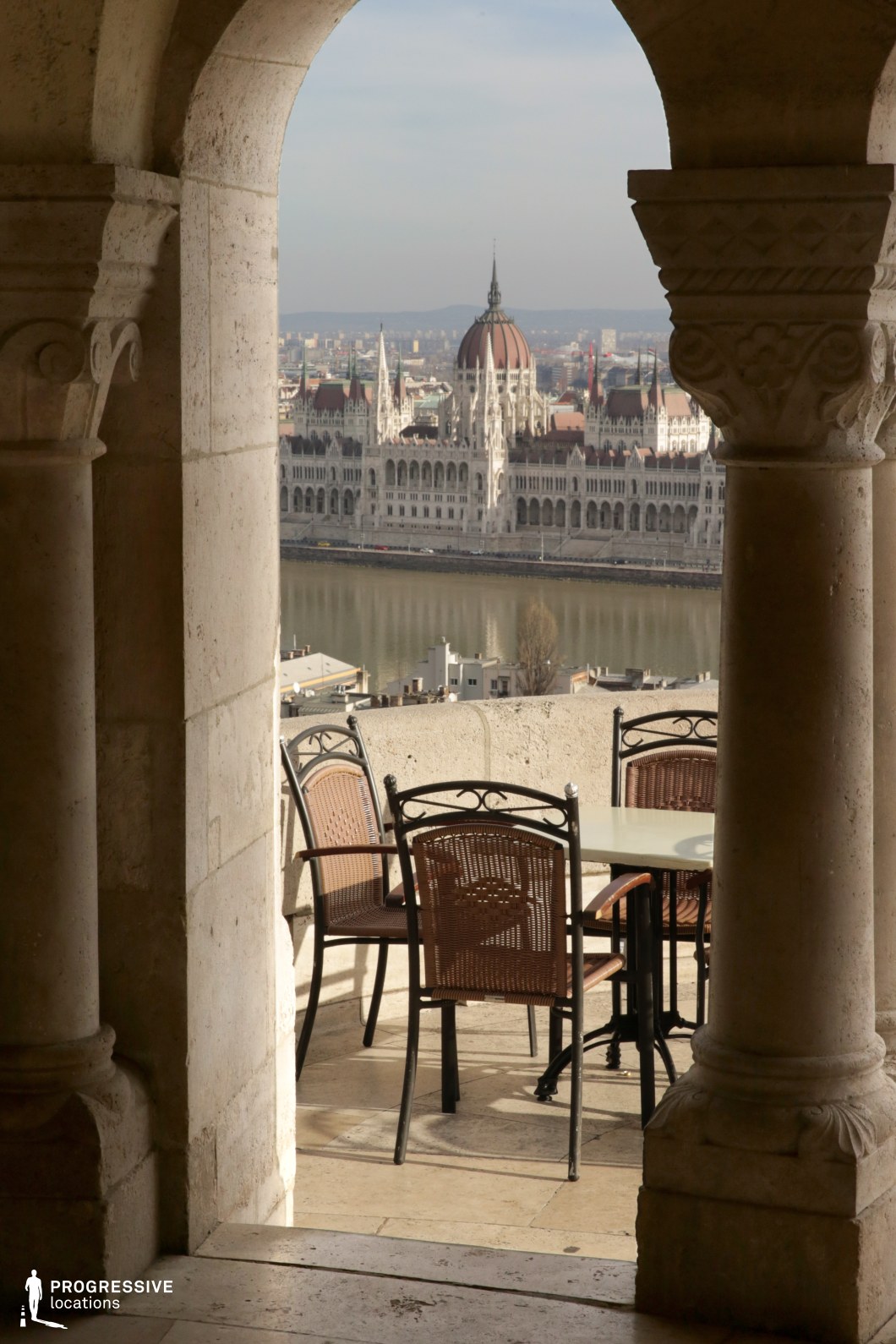 This scenic terrace offers a breathtaking panoramic view of the city, framed by elegantly carved stone archways that add a historic and architectural charm to the setting. The outdoor seating area, featuring wrought iron and wicker chairs with a simple café table, provides a picturesque spot overlooking the iconic parliament building and the river. The warm tones of the stone columns and flooring blend harmoniously with the natural light, creating a serene and timeless ambiance. This location is ideal for capturing historic European elegance, refined outdoor dining, or cinematic moments with an unparalleled cityscape backdrop.