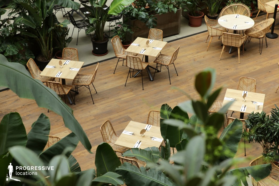 This serene dining space blends natural elements with modern simplicity, featuring wooden tables and woven rattan chairs arranged on a warm-toned wood floor. Lush tropical plants surround the seating area, creating a fresh and inviting atmosphere reminiscent of an indoor garden oasis. The combination of organic materials, soft natural lighting, and a minimalist aesthetic enhances the relaxed yet stylish ambiance. The mix of square and round tables offers versatility, making this an ideal setting for nature-inspired dining experiences, eco-conscious venues, or tranquil social gatherings.