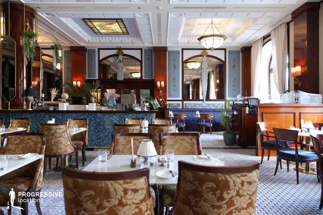 This refined restaurant blends Art Deco influences with classic European elegance, featuring a harmonious mix of soft blue accents, polished wood paneling, and intricate decorative details. The seating includes plush upholstered chairs with gold damask patterns, paired with sleek white marble tables that enhance the sophisticated ambiance. A mosaic-tiled bar with rich textures serves as a focal point, while large mirrors and arched wooden frames add depth and symmetry to the space. Stained glass ceiling panels, warm pendant lighting, and lush greenery contribute to the inviting yet upscale atmosphere, making this venue ideal for refined dining experiences, vintage-inspired settings, or high-end hospitality environments.