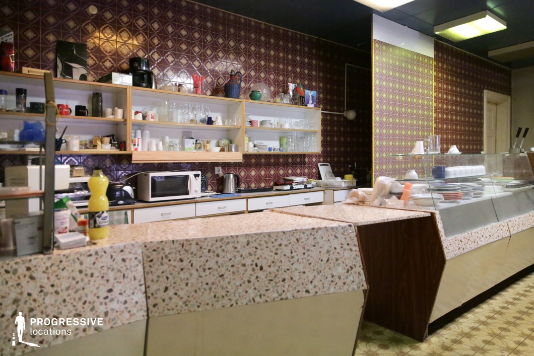 This nostalgic canteen space features a distinctly retro aesthetic with patterned ceramic wall tiles in deep burgundy tones, open shelving lined with everyday kitchen essentials, and terrazzo countertops that add a mid-century charm. The combination of wood, laminate, and metal details creates a functional yet timeworn atmosphere, reminiscent of classic cafeterias. The service counter, equipped with glass display cases, enhances the authentic vintage feel, while the patterned linoleum flooring complements the overall design. This setting is ideal for capturing a retro dining ambiance, period-piece visuals, or scenes evoking a nostalgic, everyday charm.