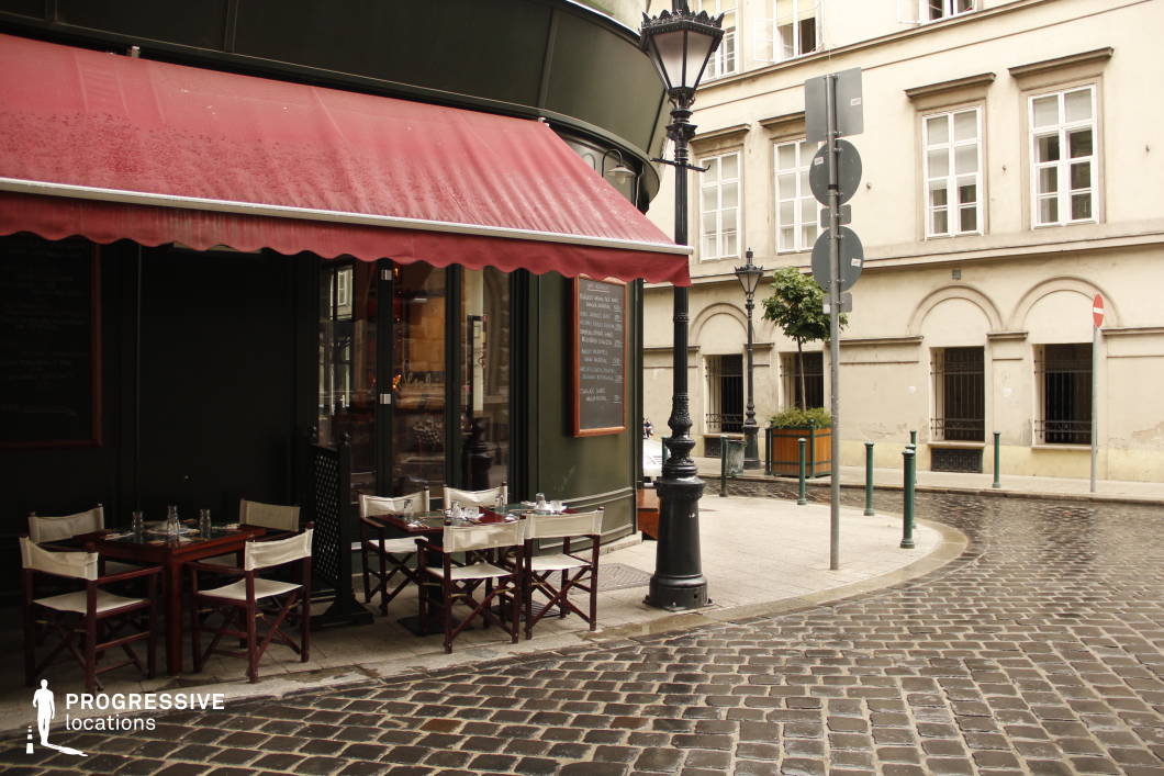 This picturesque corner café exudes European charm, featuring a deep green façade with a red awning that shelters a small outdoor seating area. Wooden tables and foldable director-style chairs line the cobblestone street, offering a cozy and intimate setting. A chalkboard menu mounted beside the entrance enhances the traditional ambiance, while ornate black street lamps and historic architecture in the background complete the classic old-town aesthetic. The combination of rich textures, muted tones, and inviting café-style elements makes this location ideal for romantic street-side dining, classic European cityscapes, or timeless urban storytelling.
