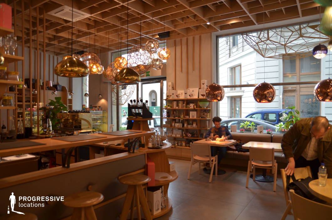 This contemporary artisan café blends Scandinavian-inspired minimalism with warm, natural materials, featuring a wooden bar counter, open shelving, and soft neutral tones. The ceiling design incorporates a geometric wooden grid, complemented by an array of pendant lights in gold, copper, and wireframe designs that create a cozy, ambient glow. The seating includes a mix of wooden stools, cushioned benches, and light-toned chairs, contributing to a relaxed yet stylish atmosphere. Large windows allow for natural light and a connection to the surrounding city, while curated decorative elements and greenery add to the welcoming environment. This setting is ideal for stylish urban cafés, modern hospitality concepts, or casual social spaces with a refined yet inviting aesthetic.