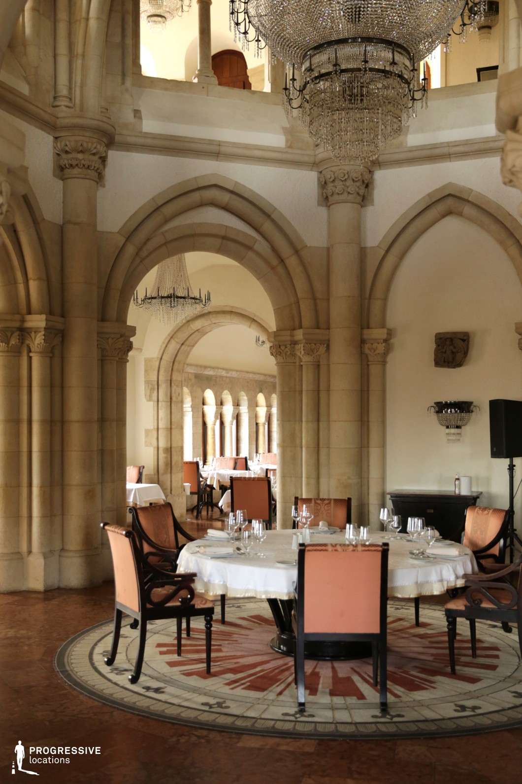 This stately dining hall showcases an exquisite blend of medieval and classical influences, featuring soaring Gothic-style stone arches, intricate column detailing, and grand chandeliers that enhance its regal ambiance. The space is defined by warm, neutral stone tones, polished terracotta flooring, and elegant round tables draped in crisp white linens, complemented by upholstered chairs with dark wood accents. Large arched openings lead to an adjoining colonnade, creating depth and a sense of historical grandeur. This setting is ideal for refined dining, historical-themed productions, or luxurious hospitality experiences that evoke a sense of timeless European elegance.