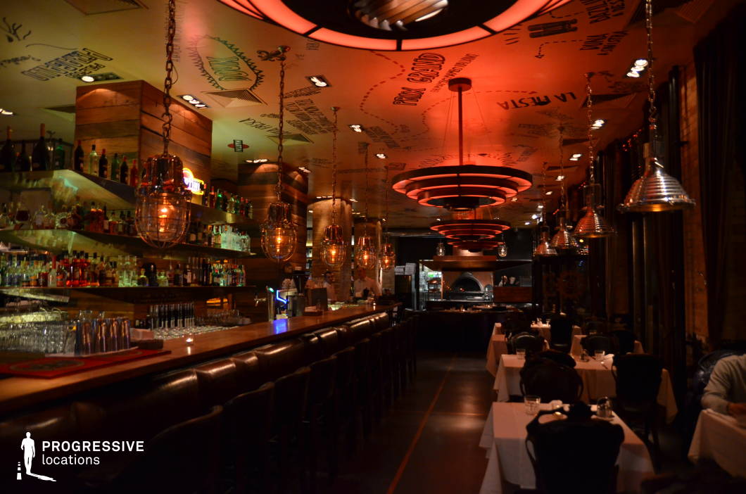 This upscale bar and restaurant feature a dynamic fusion of industrial and contemporary design elements, creating an intimate yet vibrant atmosphere. The long wooden bar is lined with plush leather stools and illuminated by hanging metal-caged pendant lights, while backlit liquor shelves enhance the warm glow. The ceiling is adorned with playful hand-drawn typography and striking circular red light fixtures that add a dramatic focal point. The open kitchen in the background, complete with a wood-fired oven, contributes to an immersive dining experience. Dark tones, exposed brick, and mixed materials make this space ideal for sophisticated nightlife scenes, high-end social gatherings, or stylish urban dining settings.