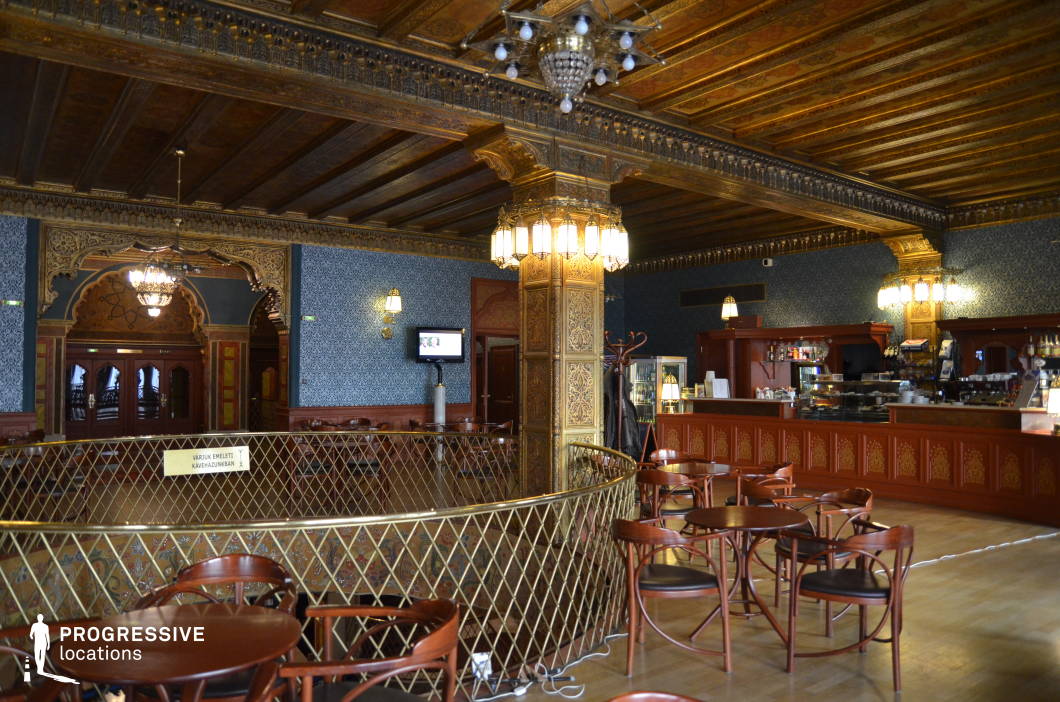 This richly decorated café exudes an old-world charm with its intricate wooden ceiling, gilded columns, and detailed Moorish-style arches. The deep blue patterned wallpaper contrasts with the warm wooden tones, while vintage chandeliers and wall-mounted lamps cast a soft, atmospheric glow. The grand counter, adorned with carved wood panels, serves as a focal point, adding to the café’s historical ambiance. The open balcony railing and classic bentwood chairs enhance the refined yet inviting setting, making it ideal for period film scenes, vintage-themed productions, or an upscale café environment with a timeless aesthetic.