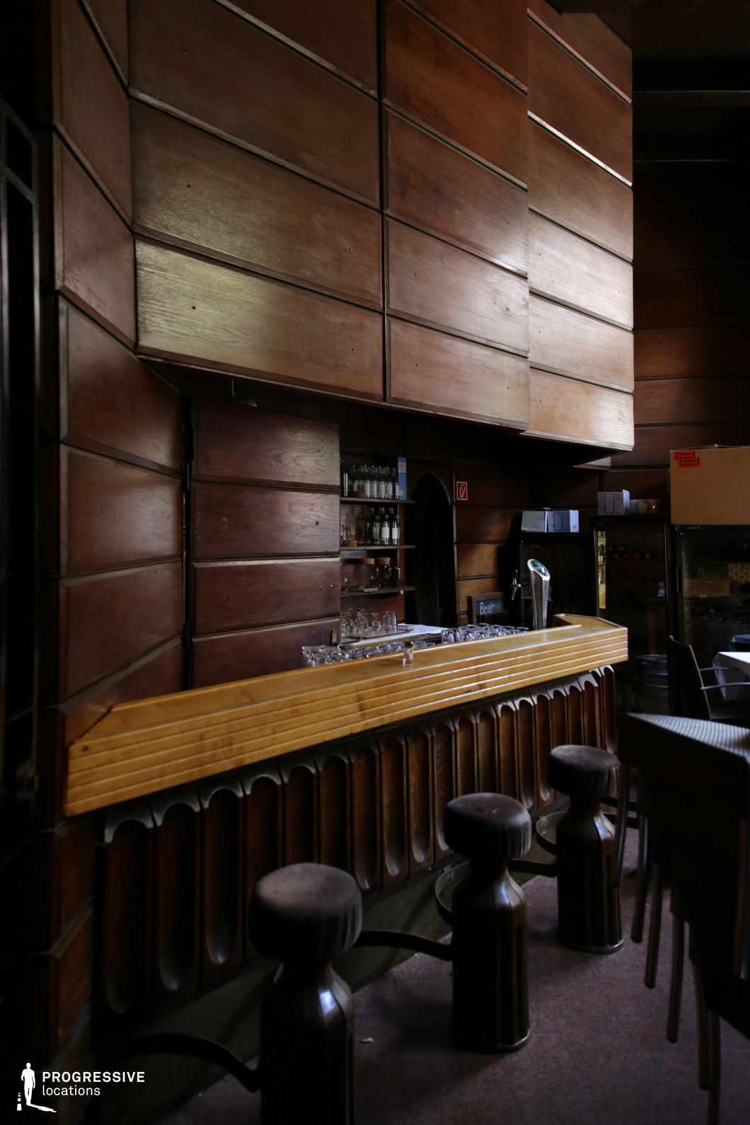 This atmospheric retro bar features dark wooden paneling with geometric patterns, evoking a mid-century modern aesthetic. The sculptural wooden stools and curved bar design add a unique, handcrafted feel, while the dim lighting enhances the intimate and nostalgic ambiance. The natural wood tones create a warm yet moody setting, ideal for vintage-themed productions, underground bar scenes, or a secluded speakeasy atmosphere. The minimal decor and integrated shelving keep the focus on the bar’s architectural details, making it a visually striking backdrop for cinematic storytelling or high-end editorial photography.